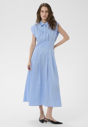 Žena s vlnitými vlasmi, oblečená v svetlomodré midi šaty bez rukávov a čierne slingback lodičky s nízkym opätkom, stojaca s jednou rukou v bok.