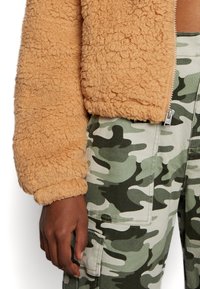 Veste zippée orange en peluche assortie à un pantalon cargo camouflé vert. La veste présente un tissu texturé ; le pantalon est doté de poches et d'un motif camouflage.