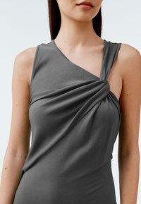 Szary top bez rękawów z zakręconym zdobieniem na ramieniu, pokazujący gładką, miękką fakturę i drapowaną tkaninę na obszarze klatki piersiowej.