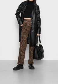 Cappotto lungo in pelle nera sopra un crop top nero, abbinato a pantaloni a strisce zebra marroni e stivali neri chunky; borsa nera in mano.
