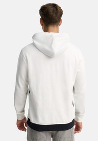 Hættetrøje i hvid med en tekstureret stof, featuring en marineblå nederste kant og sideskår. Synlig søm bagpå.