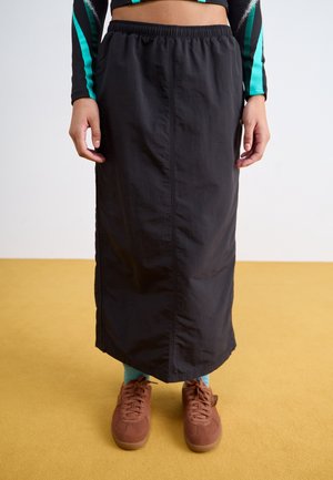 Persona con una falda larga negra, calcetines turquesa, zapatillas marrones y un top negro corto con rayas turquesas, de pie sobre un suelo amarillo.
