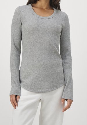 Kobieta w dopasowanej, długorękawowej, szarej prążkowanej bluzce z okrągłym dekoltem i białych spodniach, stojąca na jednolitym tle.