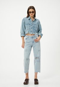 Camisa de mezclilla recortada con dos bolsillos en el pecho, color azul claro, con dobladillo deshilachado. Vaqueros azul claro desgastados con dobladillos desiguales y rasgaduras. Zuecos de plataforma marrones.