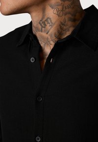 Schwarzes Hemd mit Kn opfverschluss und weicher Textur. Verfügt über einen klassischen Kragen, sichtbare Knöpfe und eine Nahaufnahme von Nackentattoos.