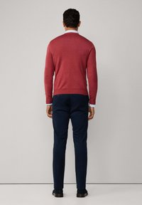 Czerwona sweterka dziergana z długimi rękawami i detalami mankietów, noszona na koszulę z kołnierzykiem, w połączeniu z granatowymi spodniami w wąskim kroju. Spodnie mają gładką teksturę.