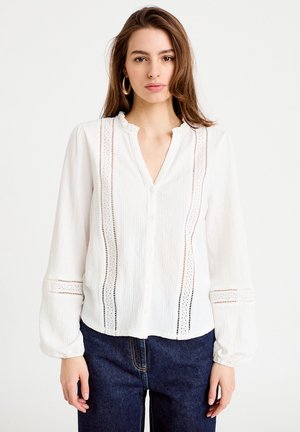 Biała bluzka z długim rękawem, z falbankowym kołnierzem, zapinana z przodu na guziki, z pionowymi wzorami dziurek po bokach, zestawiona z ciemnymi jeansami.