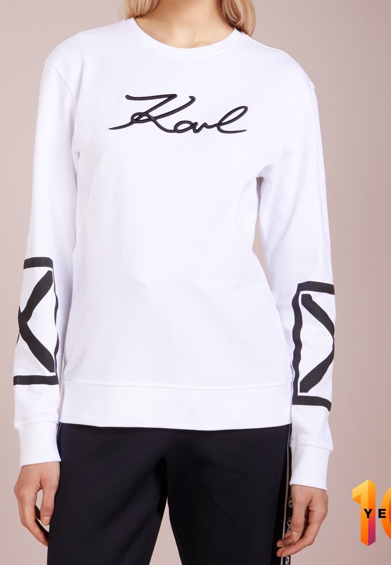 Personne portant un sweat-shirt blanc à manches longues avec l'inscription "Karl" sur la poitrine et des motifs géométriques noirs sur les manches, associé à un pantalon noir.