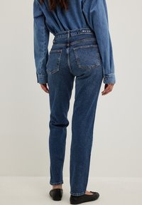 Jeans in denim blu con vita alta e design a cinque tasche. Presentano un lavaggio sbiadito e dettagli cuciti lungo le cuciture. Indossati con una camicia blu.