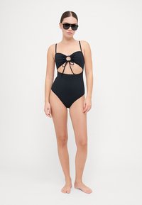 Maillot de bain une pièce noir avec une découpe en trou de serrure, des bretelles réglables et un accent noué au niveau de la poitrine. Texture de tissu lisse et extensible.