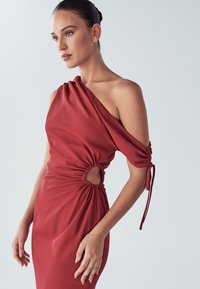 Sarkanā kleita ar vienu plecu, ar saplēstu audumu, griezuma detaļu viduklī un regulējamām piedurknēm. Gluda tekstūra ar pieguļošu siluetu.