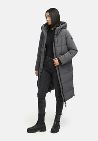 Lång, grå puffjacka med huva, kviltad textur, frontdragkedja, sidofickor, bärs över en svart topp och svarta jeans, parat med svarta stövlar.