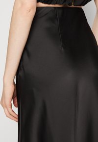 Gros plan sur la main d'une personne reposant à côté de sa hanche, portant une jupe noire en satin lisse taille haute et un top noir court.