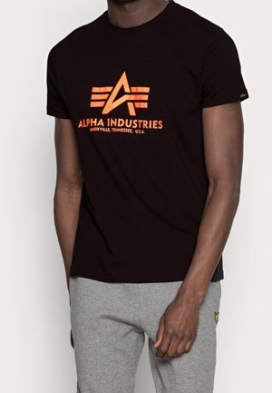 Homme portant un t-shirt noir Alpha Industries avec un logo et un texte orange, assorti à un pantalon de survêtement gris clair, debout devant un fond uni.