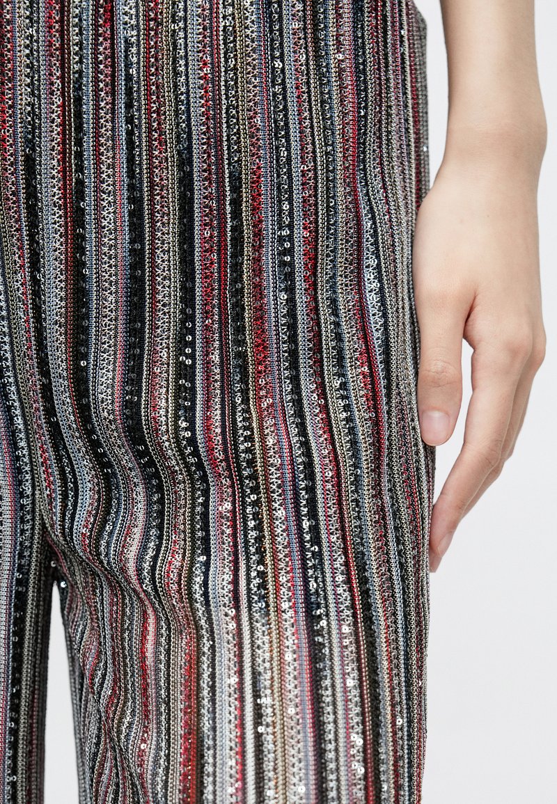 Multifarvede palietbukser med lodrette striber i nuancer af rød, sort, sølv og guld. Nærbillede af tekstur og håndplacering.