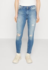 Blue denim skinny jeans with distressed rips on the knees, light wash, five-pocket design, paired with beige sneakers.