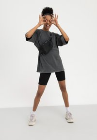 Mladić u prevelikoj sivoj majici, crnim biciklističkim kratkim hlačama, tenisicama i torbicom preko tijela koji stvara naočale rukama i smiješi se na bijeloj pozadini.