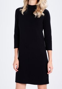 Robe noire à manches longues avec un col montant, présentant une coupe droite et une texture lisse et unie. Sans motifs ni embellissements.