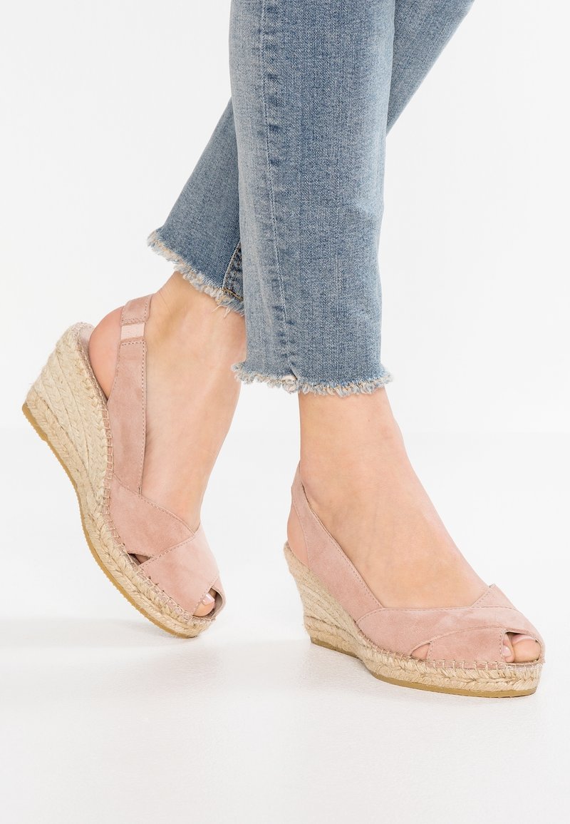 Pieds chaussés de sandales compensées en espadrilles en daim rose associées à un jean bleu clair à ourlet effiloché sur fond blanc.