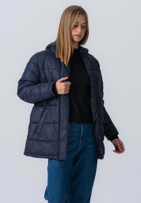 Marineblå puffjakke med hætte, quiltet struktur, front knaplukning og sidelommer. Sammen med en sort sweater og denimjeans.