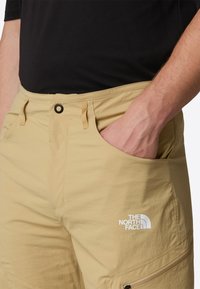 Beige cargo-housut, jotka on valmistettu kevyestä kankaasta. Niissä on etutasku, säädettävä vyötärö ja valkoinen logoerittely reidessä.