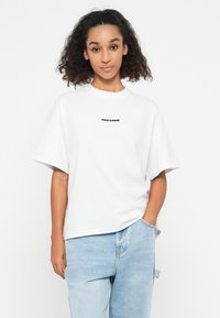 Woman with curly hair wearing a white oversized T-shirt with "PEGADOR" text and light blue jeans, standing with one hand in pocket.