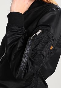 Svart bomberjacka med en glänsande, slät textur. Har ribbade mudd och krage, dragkedjeficka på ärmen och etikett med texten "TA BORT FÖRE FLYGNING".