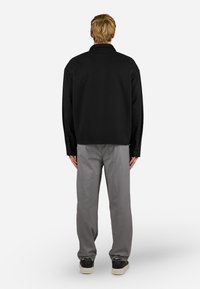 Camisa oversized preta feita de tecido, com corte reto e botões nos punhos, combinada com calças cinzas e sapatos pretos.