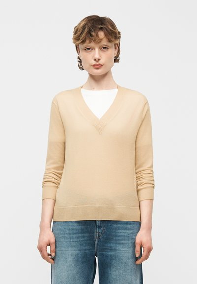 Personne aux cheveux courts portant un pull beige à col en V sur un haut blanc et un jean bleu en denim, debout devant un fond clair uni.