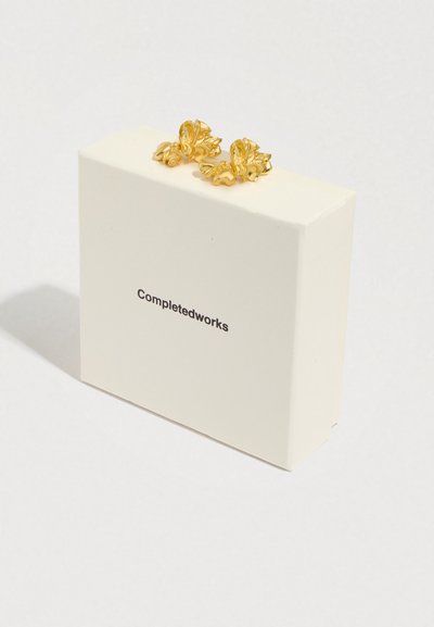 Brincos dourados em forma de flor repousam sobre uma caixa branca com a inscrição "Completedworks" a preto. O design apresenta detalhes embossados e intrincados.