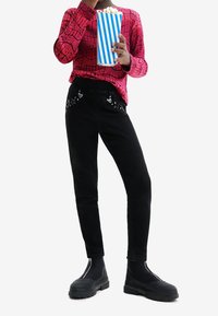 Černé manšestrové kalhoty s vyšívanými srdíčky a hvězdami, doplněné růžovým tričkem s dlouhým rukávem s potiskem a černými masivními botami, držící popcorn.