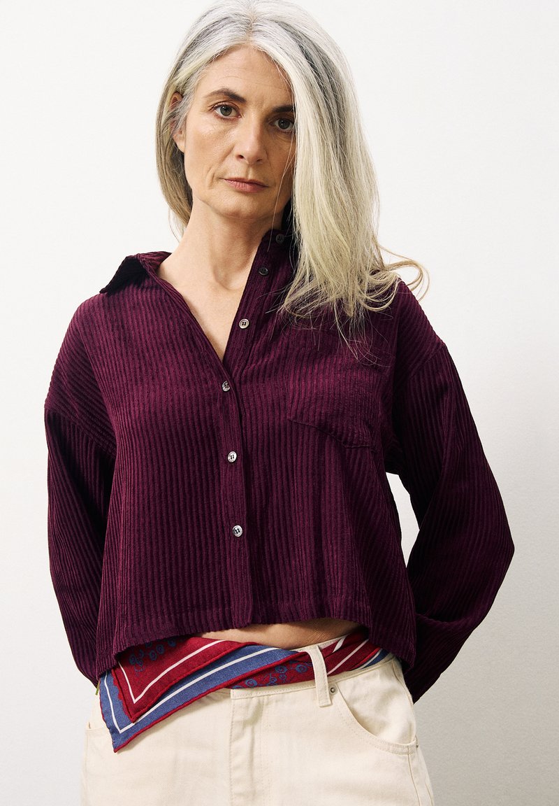 Chemise en velours côtelé bordeaux à boutons avec col, présentant une poche frontale et un design court. Un foulard à motifs est noué à la taille.