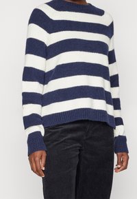 Pull marin et blanc à rayures avec un col rond, fabriqué en tissu doux. Dispose de poignets et d'un ourlet côtelés. Porté par-dessus un pantalon en velours côtelé noir.