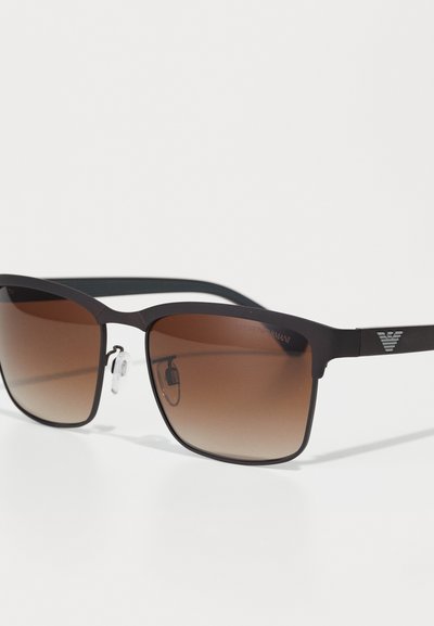 Black frame sunglasses with gradient brown lenses. Square shape, featuring clear nose pads and a silver logo detail on the arm.