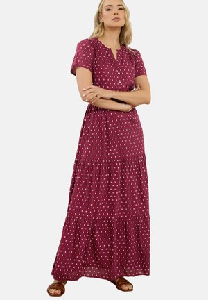 Frau steht mit verschränkten Armen, trägt ein langes burgunderrotes Kleid mit weißen senkrechten Strichmustern und braune Sandalen vor einfachem Hintergrund.