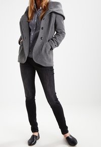 Abrigo de mezcla de lana gris con gran solapa y botones dobles, combinado con una camisa a cuadros, vaqueros oscuros y zapatos negros tipo slip-on.