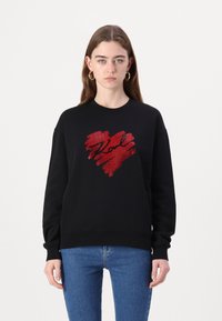 Svart sweatshirt med ett rött hjärtdesign i penseldragstyp och "Love" i svart skrivstil, parat med blå jeans.