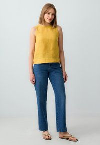Top senza maniche color giallo senape in lino abbinato a jeans dritti blu e infradito marroni, caratterizzato da un design semplice e rilassato.