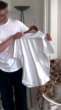 T-shirt branco de mangas curtas numa cabide de madeira, com um decote redondo e textura suave. Segurado por uma pessoa vestida de forma casual.
