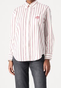 Camicia bianca con colletto a punta, righe verticali rosse e taschino sul lato sinistro ricamato con "GANT" in rosso. Realizzata in cotone, maniche lunghe.