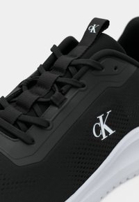 Baskets noires avec semelle blanche, lacets noirs et logo blanc "cK" sur le côté et la languette.