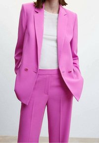 Jasnoróżowy szyty na miarę garnitur z podwójnym zapięciem, z klapami i dopasowanymi szerokimi spodniami; gładki materiał, minimalne detale.