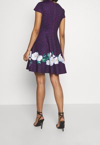 Lila klänning med leopardmönster, korta ärmar, utsvängd kjol och vita blommor i kanten. Dragkedja bak. Klackskor.