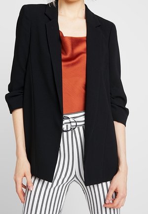 Femme portant un blazer noir sur un top en satin couleur rouille et un pantalon blanc taille haute avec des rayures verticales noires et une ceinture à double boucle.
