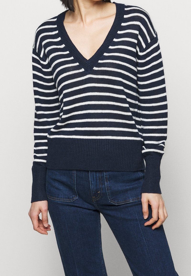 Pull rayé bleu marine et blanc avec un col en V profond, des poignets et un ourlet côtelés. Porté avec un jean bleu foncé, présentant une coupe ample et une texture tricotée.