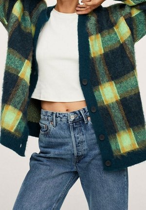 Personne portant un haut court blanc, un jean taille haute bleu et un cardigan à carreaux vert foncé avec de gros boutons.