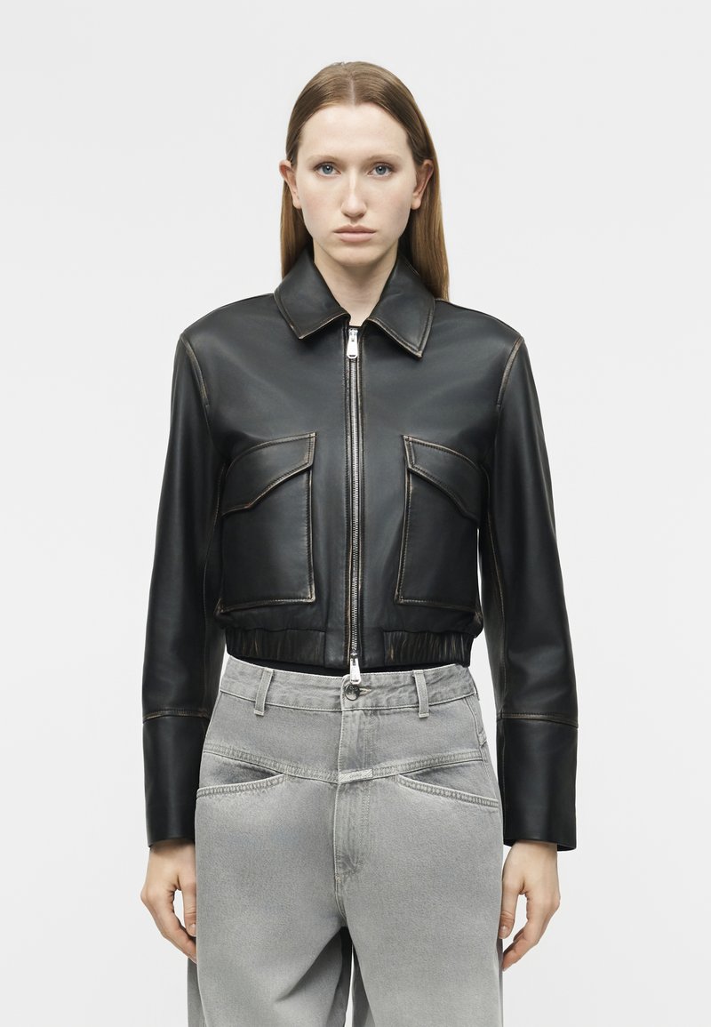 Γυναίκα με ίσια καστανά μαλλιά φοράει μαύρο δερμάτινο σακάκι με φερμουάρ και μεγάλες μπροστινές τσέπες, καθώς και ανοιχτό γκρι τζιν ψηλόμεσο παντελόνι.