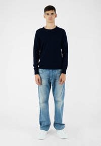 Maglione blu navy con collo rotondo, abbinato a jeans azzurri chiari con una texture sbiadita, e sneakers bianche su uno sfondo semplice.