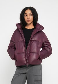 Vrouw in een donkerpaarse pufferjack, zwarte shirt en grijze cargobroek, staand met handen in de zakken tegen een effen achtergrond.
