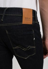 Jeans en denim bleu foncé avec une étiquette en cuir à l'arrière indiquant "REPLAY HYPERFLEX". Comprend des coutures jaunes et un design de poche unique.